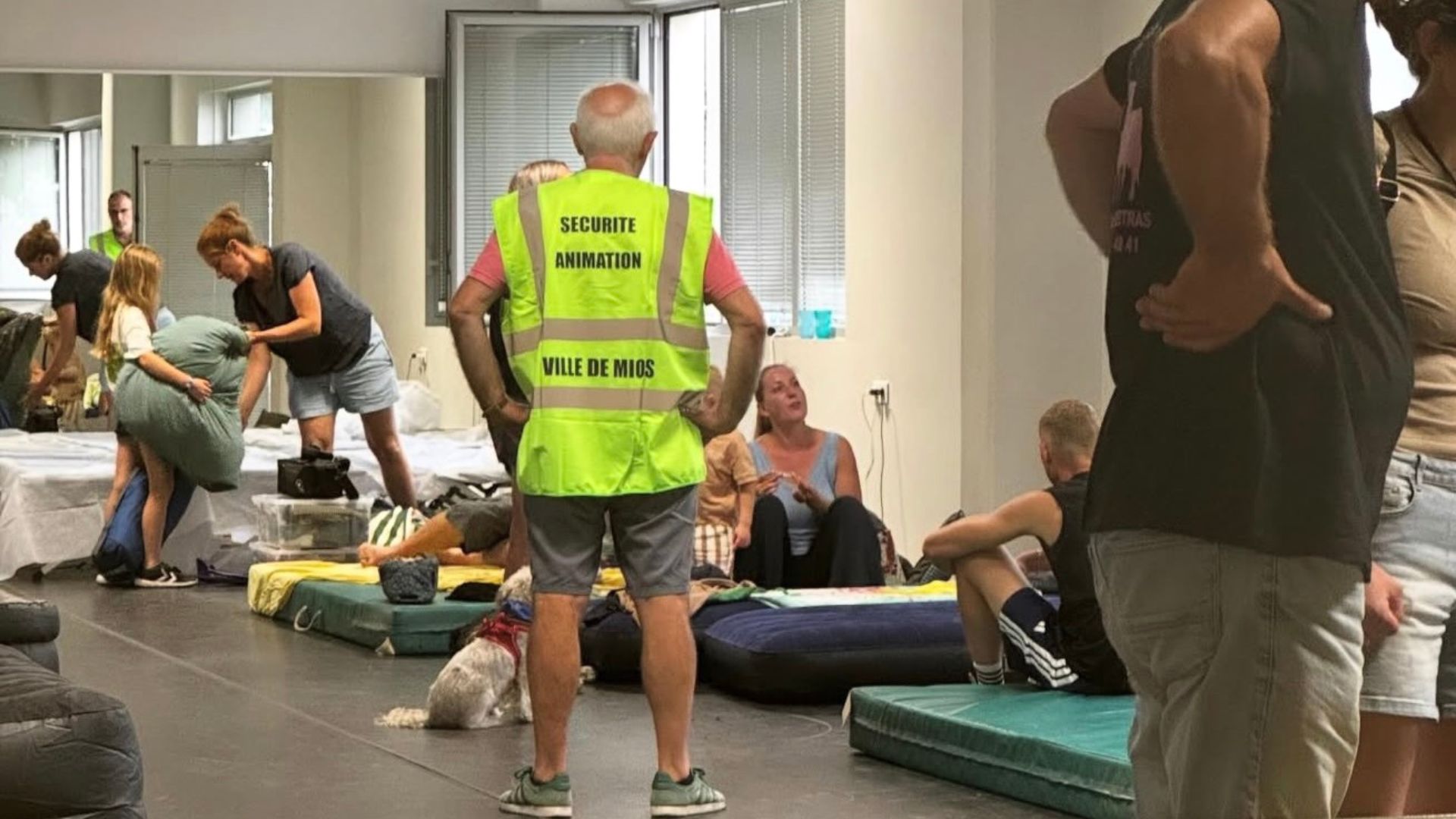 une salle rempli de personnes assises sur des matelas, au centre un homme avec un gilet jaune