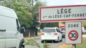des voitures dont une voitures blanche quivie d'un utilitaire blanc passent derrière le panneau : Lège-Cap Ferret