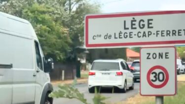 des voitures dont une voitures blanche quivie d'un utilitaire blanc passent derrière le panneau : Lège-Cap Ferret