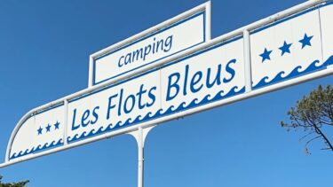 Un panneau blanc avec des écritures bleues : "camping, les flots bleus"