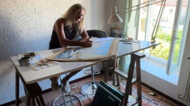 Un homme aux cheveux longs et atablé à une table de dessin. Une fenêtre illumine la pièce à sa droite