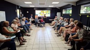 Dans une salle au mur bleus foncés des personness sont assises sur des chaises alignées contre les murs.