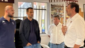 Deux hommes en chemises blanches à droite discutent avec deux hommes en pull bleu à gauche; ils sont à l'intérieur d'une salle devant une fenetre