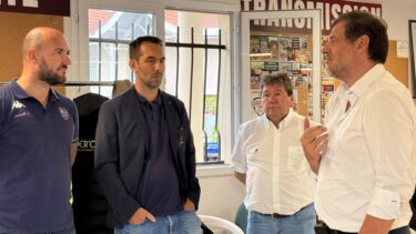 Deux hommes en chemises blanches à droite discutent avec deux hommes en pull bleu à gauche; ils sont à l'intérieur d'une salle devant une fenetre