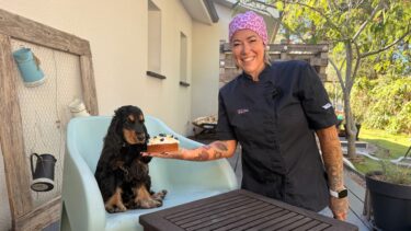 femme en tenue de pâtissière souriante qui tient un gâteau dans la main et qui pose à côté d'un cocker installé sur une chaise