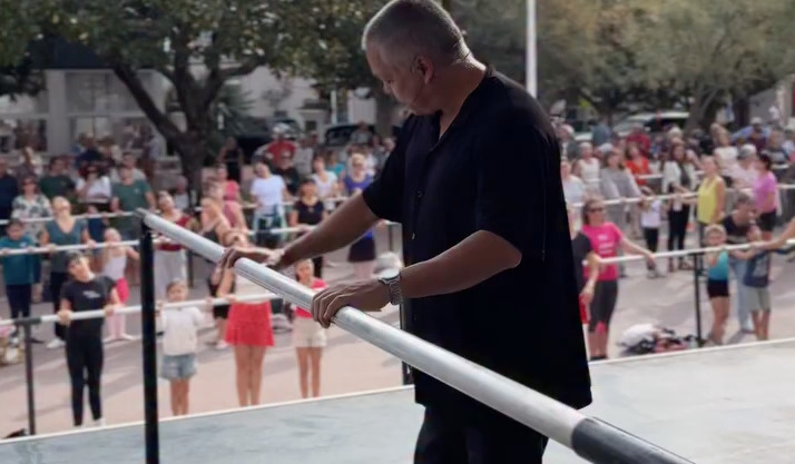Un homme au cheveux courts habillé de noir sur une scène se tient à une barre de danse classique. en contrebas, des danseurs et danseuses suivent le cours