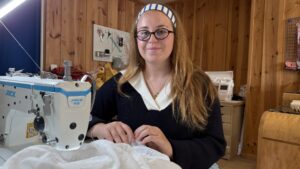 une jeune femme avec des lunettes et unbandeau dans les cheveux se tient derrière une machine a coudre. elle tient dans les mains un tissu blanc. Mur en bois derrière elle.