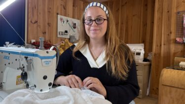 une jeune femme avec des lunettes et unbandeau dans les cheveux se tient derrière une machine a coudre. elle tient dans les mains un tissu blanc. Mur en bois derrière elle.