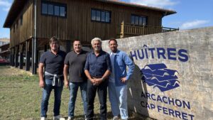 4 hommes pos een ligne devant un panneau "huitres arcachon- Cap ferret". Derrière une un grand batiment en bois