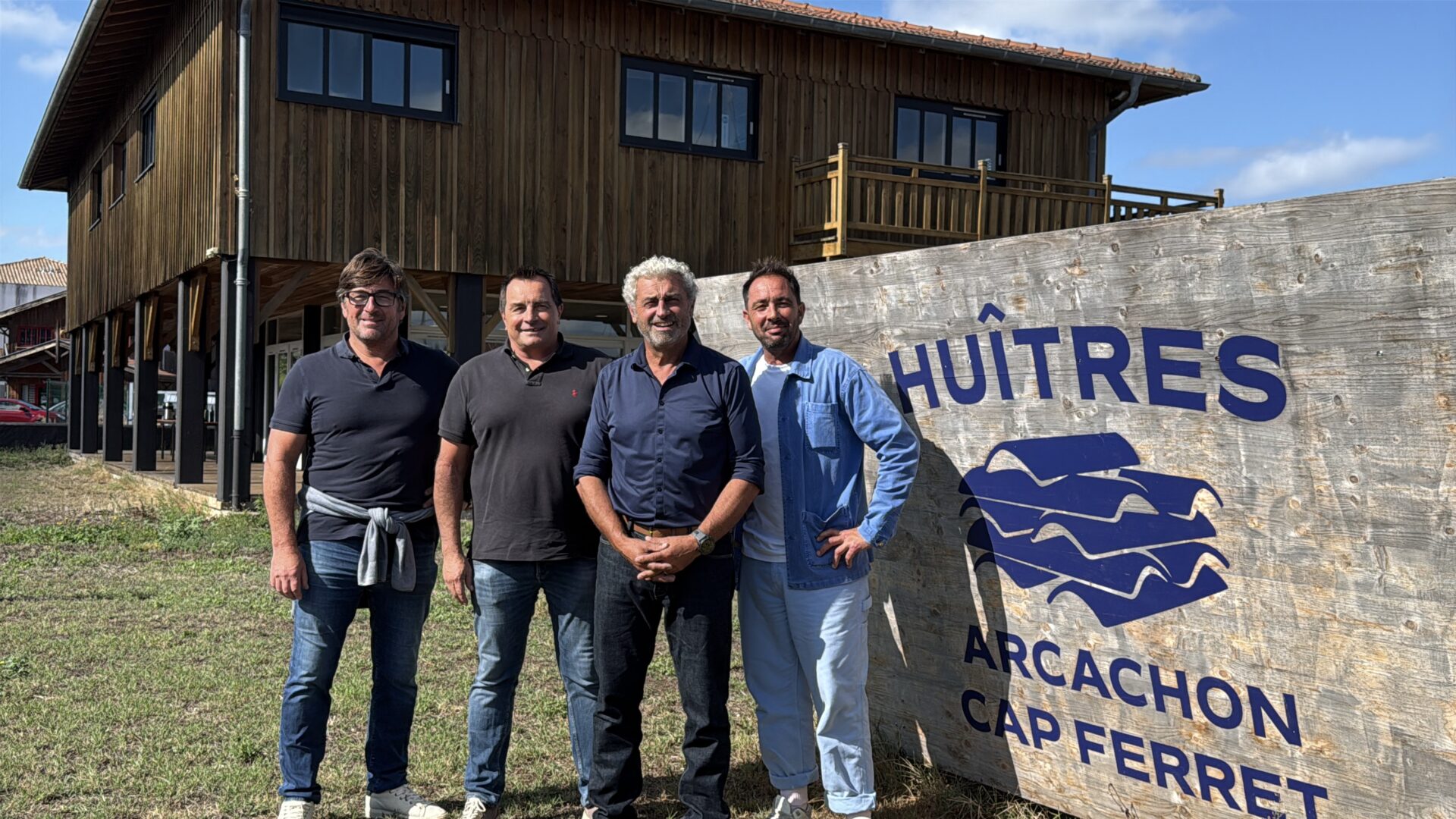 4 hommes pos een ligne devant un panneau "huitres arcachon- Cap ferret". Derrière une un grand batiment en bois