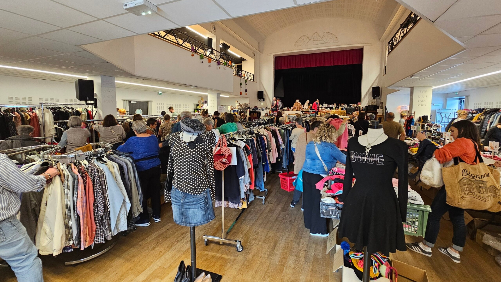 dans la salle publique du Teich, des rangées de portants avec des vêtements et des personnes qui participent à la braderie
