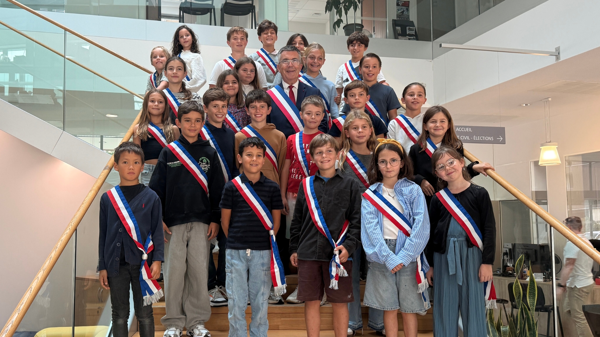 25 jeunes enfants et un adulte au milieu installés sur l'escalier de l'accueil de la mairie de la teste. Ils portent des écharpes tricolores.