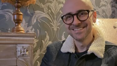 Un homme chauve avec des lunette fait un sourire devant un papier peint baroque et une lampe à frange vintage